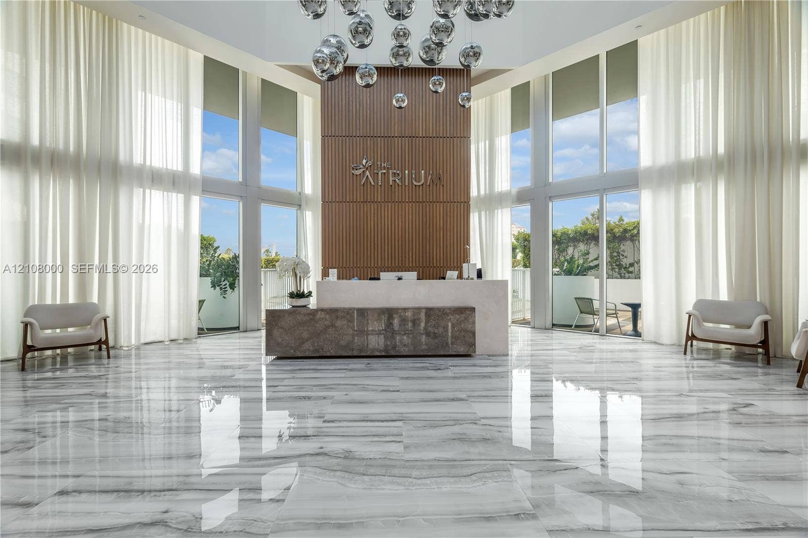 Aventura, FL 33180 Double-height ceiling, floor-to-ceiling windows, polished stone flooring, custom vertical wood-slat feature wall, and tiered spherical pendant lighting