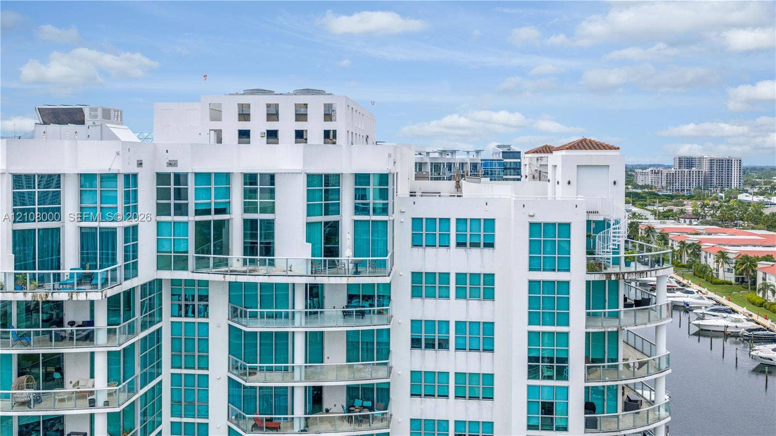 Aventura, FL 33180 High-rise exterior featuring a white stucco facade, floor-to-ceiling glass paneling, multiple levels of private glass-railed balconies, and a marina-front location