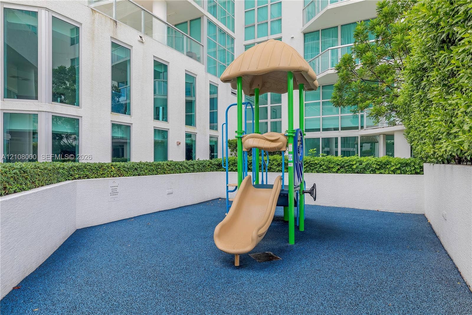 Aventura, FL 33180 Outdoor recreational area featuring a structured play system with a slide, resilient rubberized flooring, and perimeter privacy hedges
