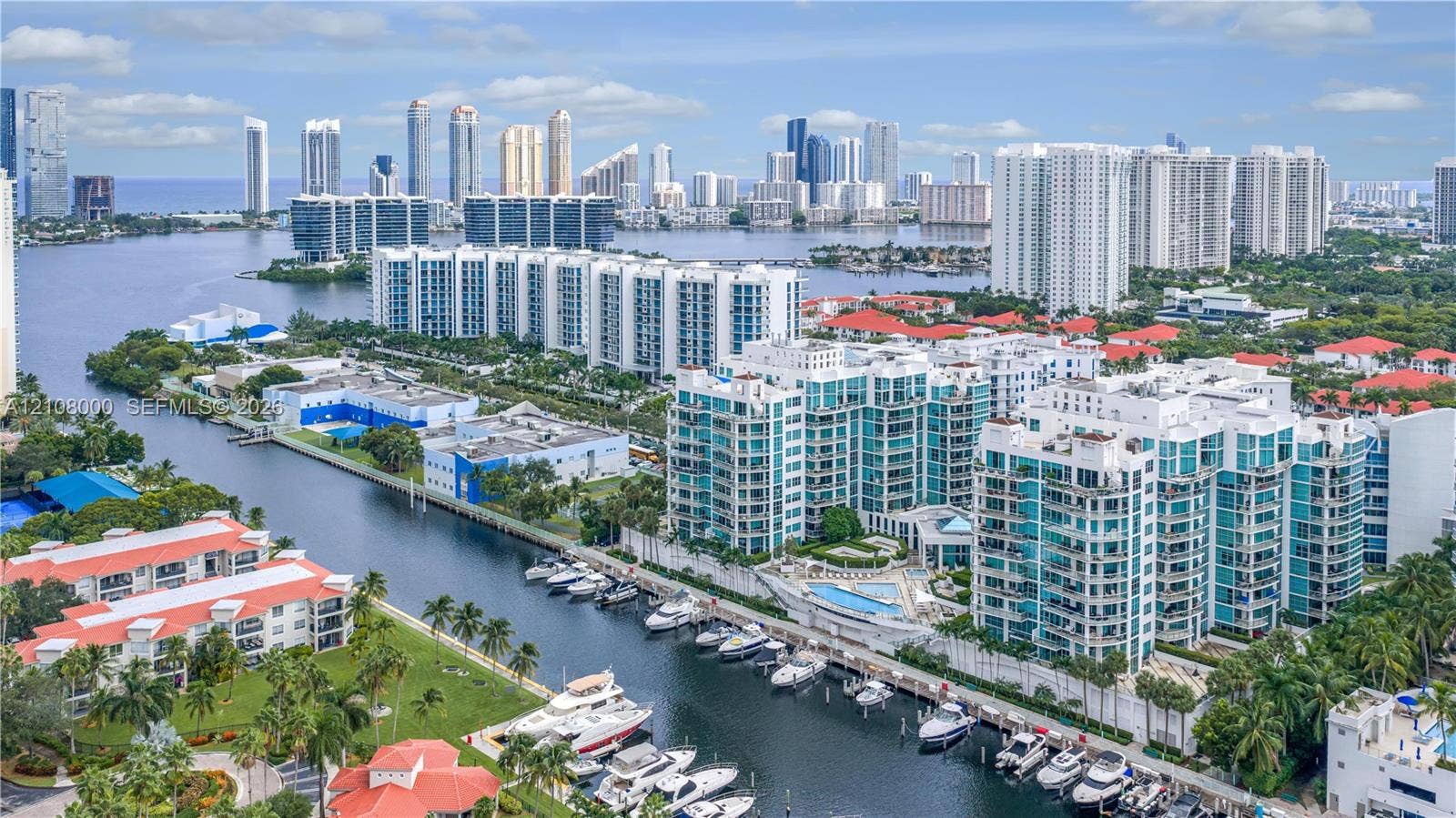 Aventura, FL 33180 Aerial perspective of a multi-story residential complex featuring waterfront access, private boat slips, extensive hardscaping, and a swimming pool, situated against an urban skyline backdrop