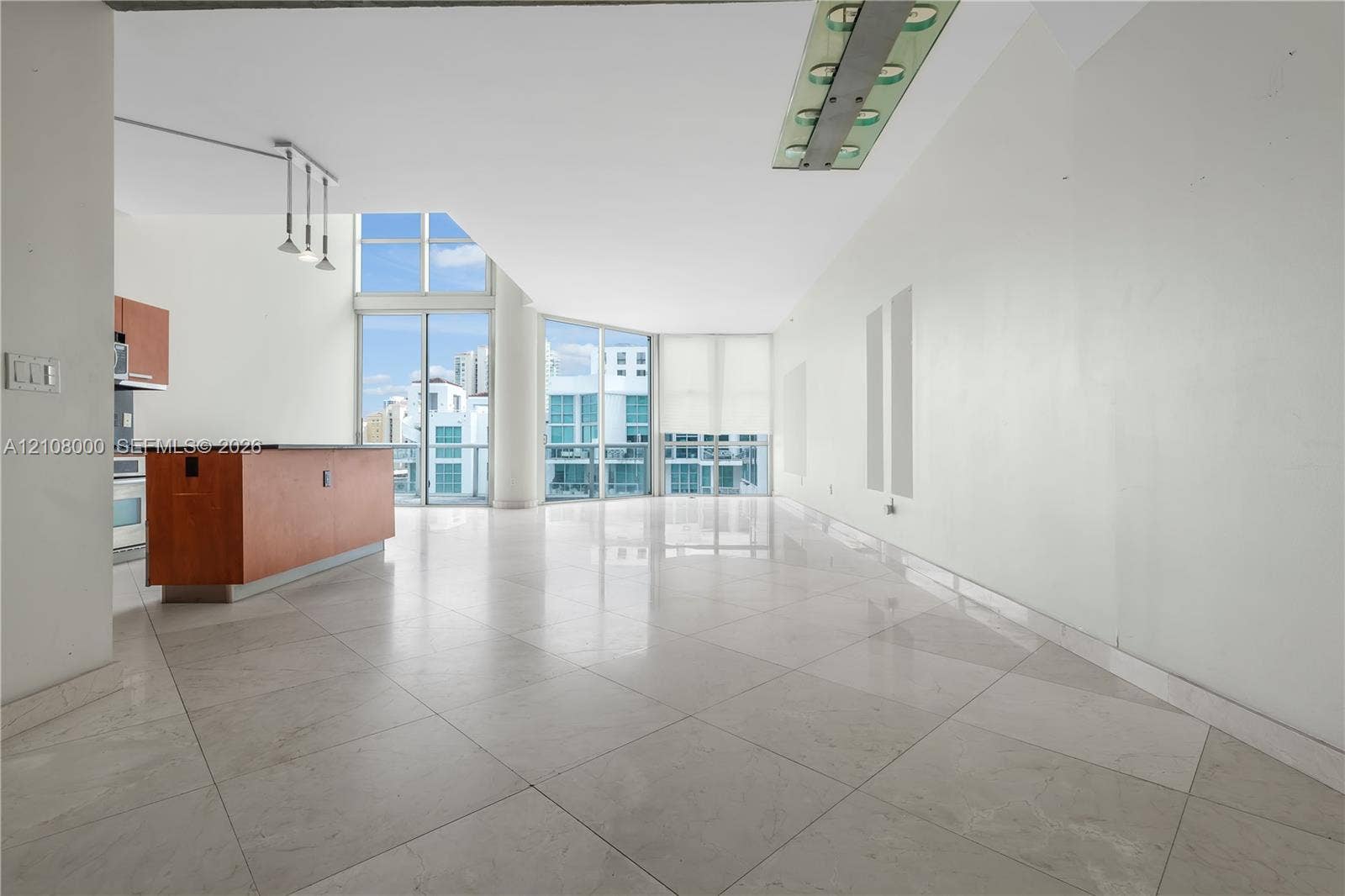 Aventura, FL 33180 Open living area featuring large-format stone-finish flooring, floor-to-ceiling windows, a center kitchen island with dark surfacing, and contemporary recessed lighting