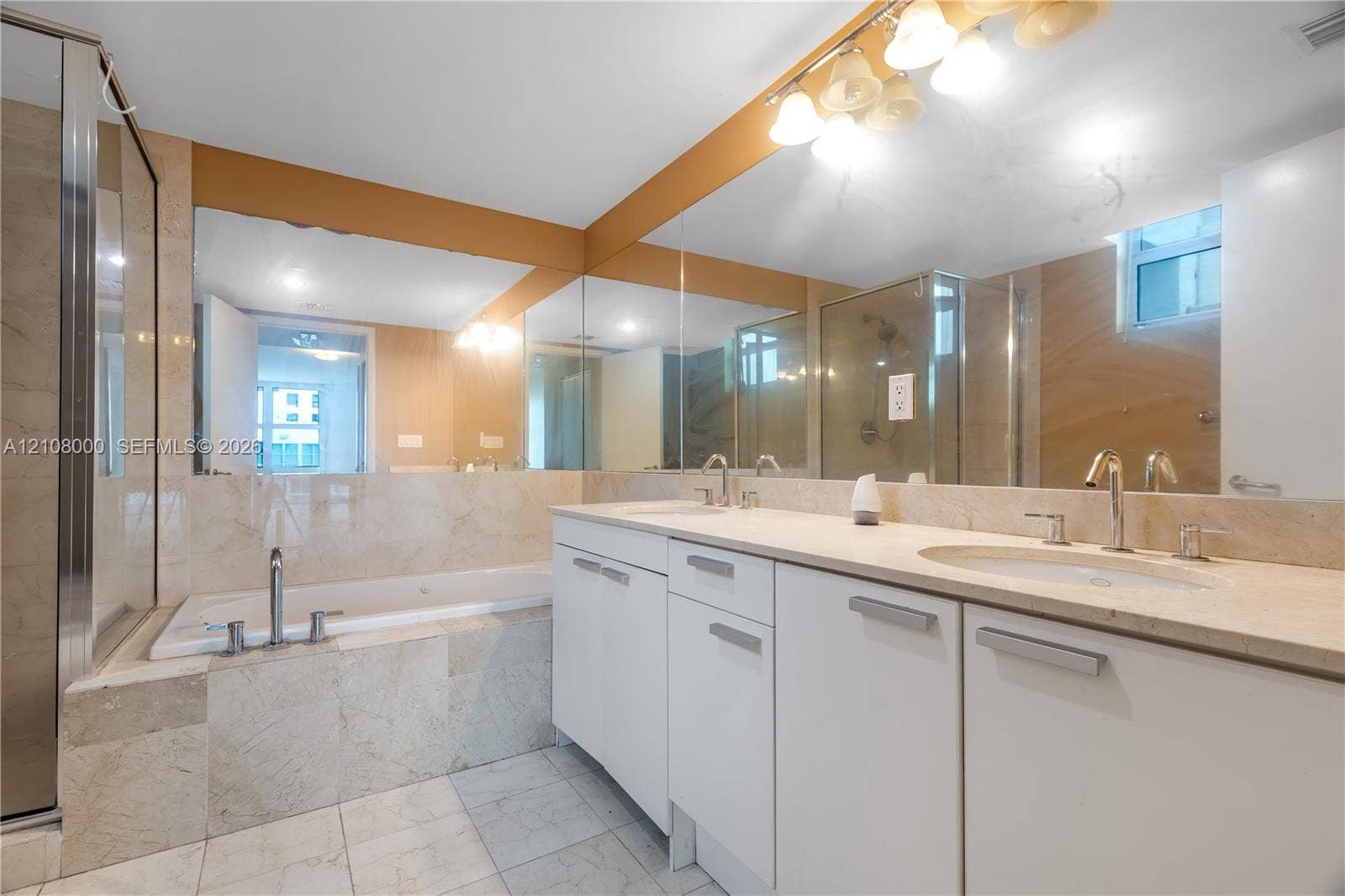 Aventura, FL 33180 Bathroom featuring a stone-surround soaking tub, dual-sink vanity with light-toned countertop, full-width wall mirror, and a glass-enclosed shower