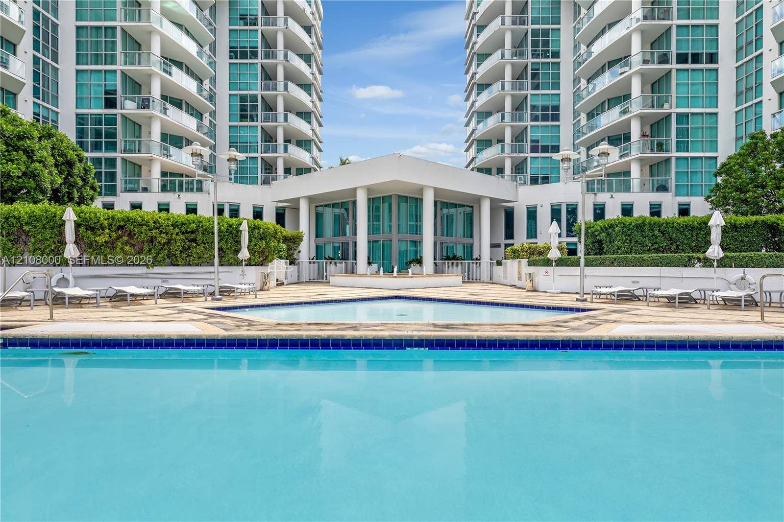 Aventura, FL 33180 Swimming pool with decorative tile border, expansive stone-paver sun deck, manicured hedges, and multi-story high-rise architecture with glass-paneled balconies