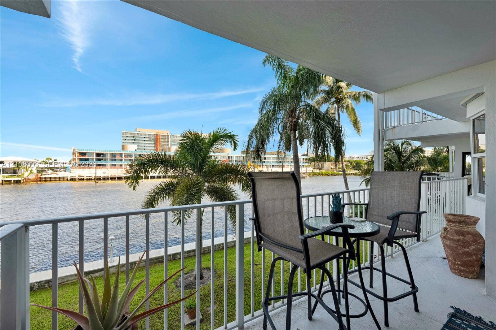 1536 SE 15th Court, #207, Deerfield Beach, FL 33441 Covered balcony featuring white vertical railing and concrete flooring, overlooking the waterfront with mature palm trees and manicured lawn