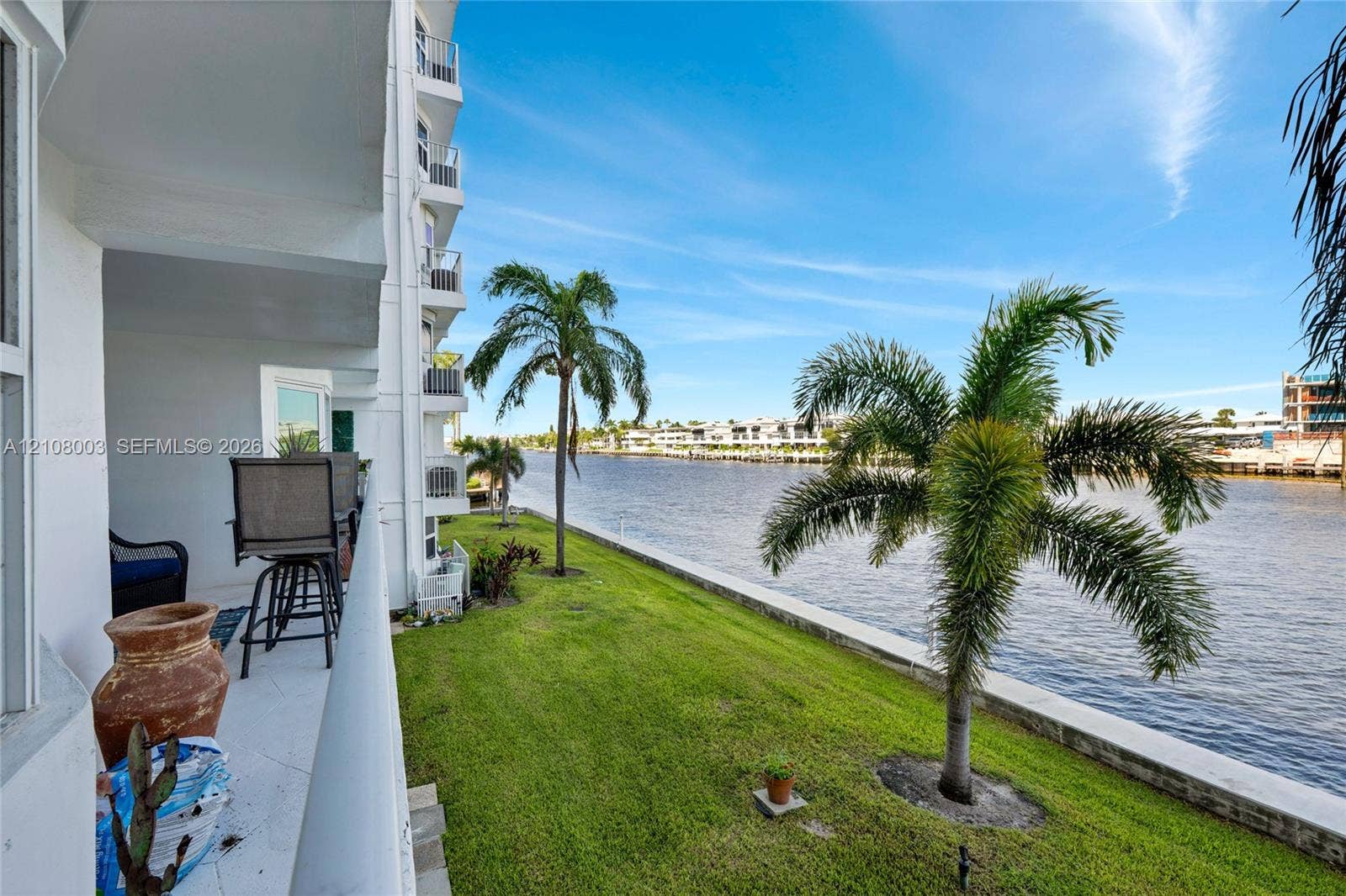 1536 SE 15th Court, #207, Deerfield Beach, FL 33441 Covered balcony featuring white tiled flooring and a metal railing, overlooking a waterfront lawn with palm trees and a concrete bulkhead