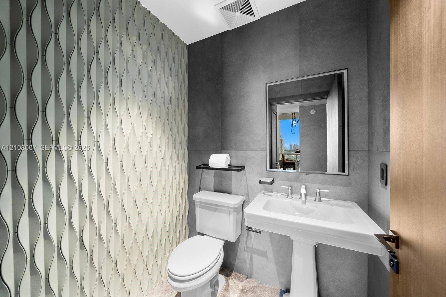 Textured dimensional wall tile, matte charcoal large-format wall surfacing, pedestal sink with polished chrome hardware, and wood-finish door