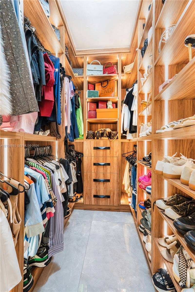 Custom walk-in closet featuring floor-to-ceiling wood-finish shelving, integrated vertical LED strip lighting, a central four-drawer dresser with matte black hardware, and large-format tile flooring