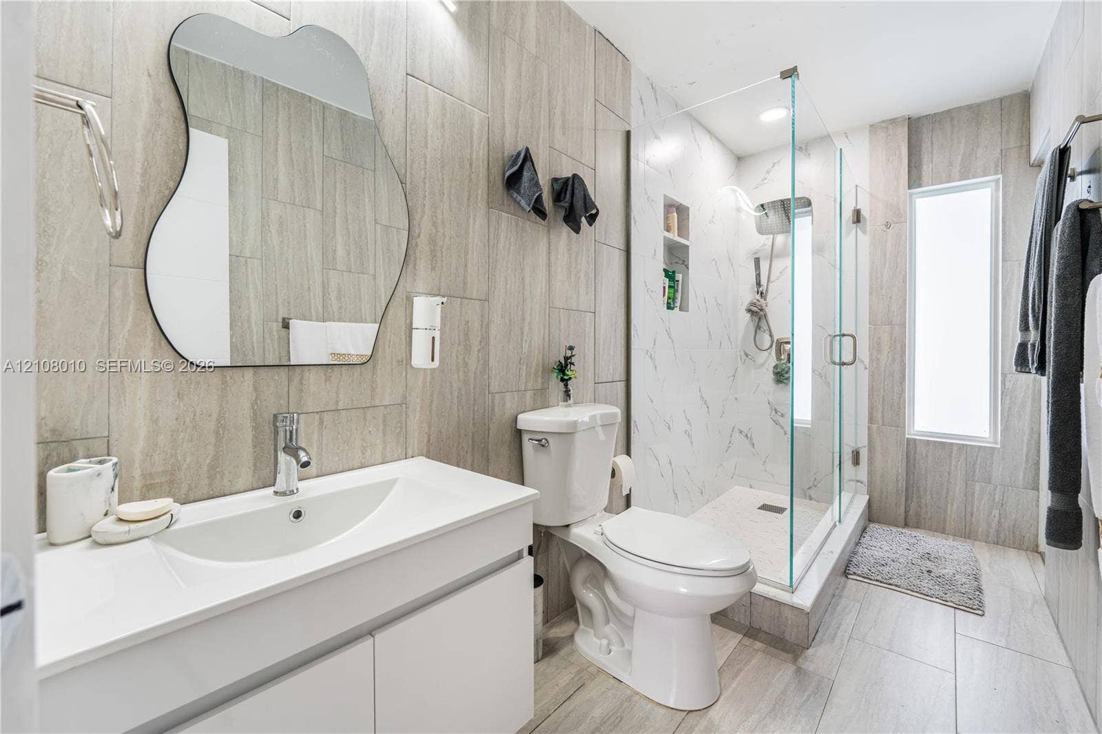 Bathroom featuring vertical wood-finish wall tile, white vanity with an integrated sink, frameless glass-enclosed shower with a recessed shelf, and an irregular-shaped wall mirror
