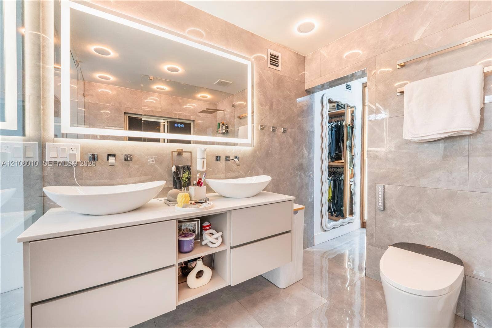 Floating dual-sink vanity with matte white vessel basins, large wall-to-wall LED-backlit mirror, and polished stone-finish tile flooring and wall surfaces