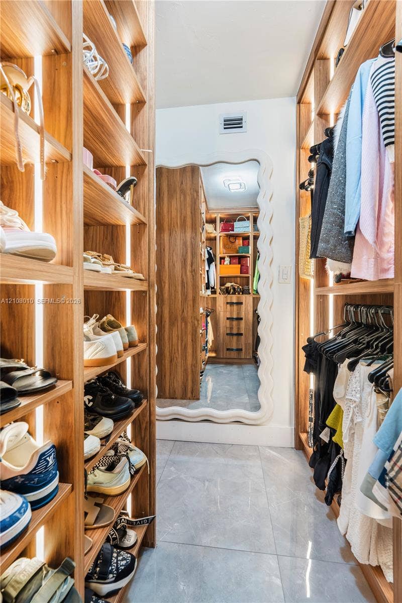 Walk-in closet featuring custom wood-finish shelving with integrated vertical lighting, polished tile flooring, and a full-length mirror with a decorative wave-edge frame
