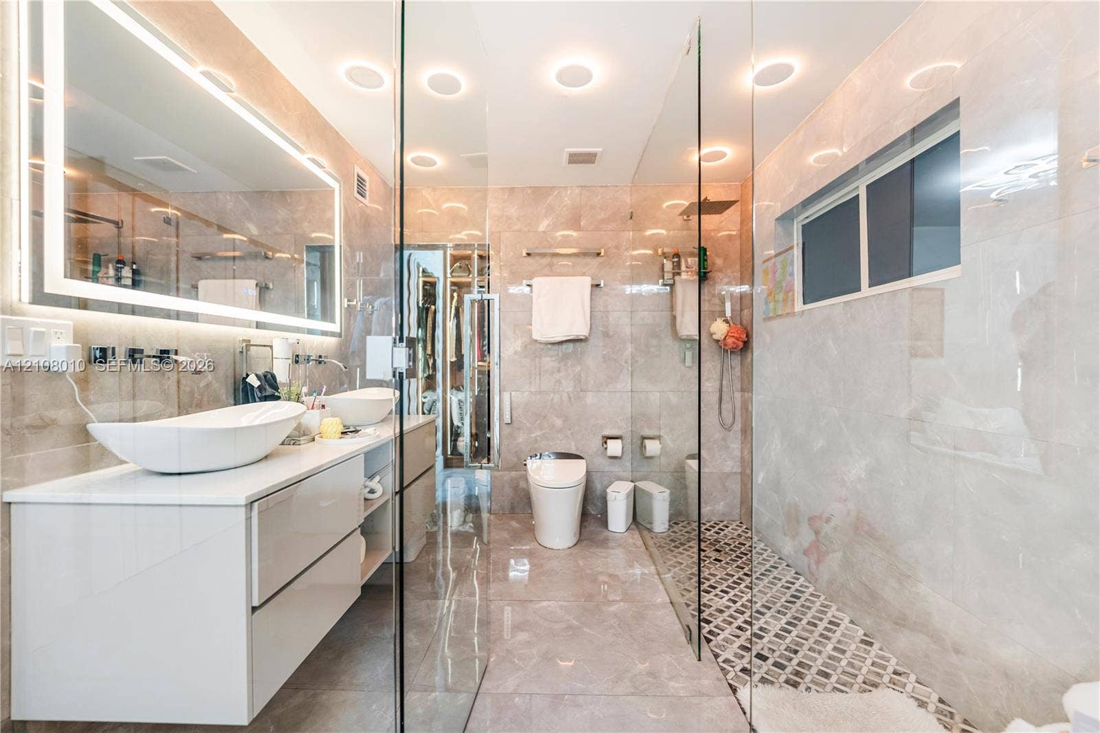 Modern dual-basin floating vanity with vessel sinks, backlit wall-mounted mirror, walk-in glass-enclosed shower, floor-to-ceiling stone-tiled walls, and recessed ceiling lighting