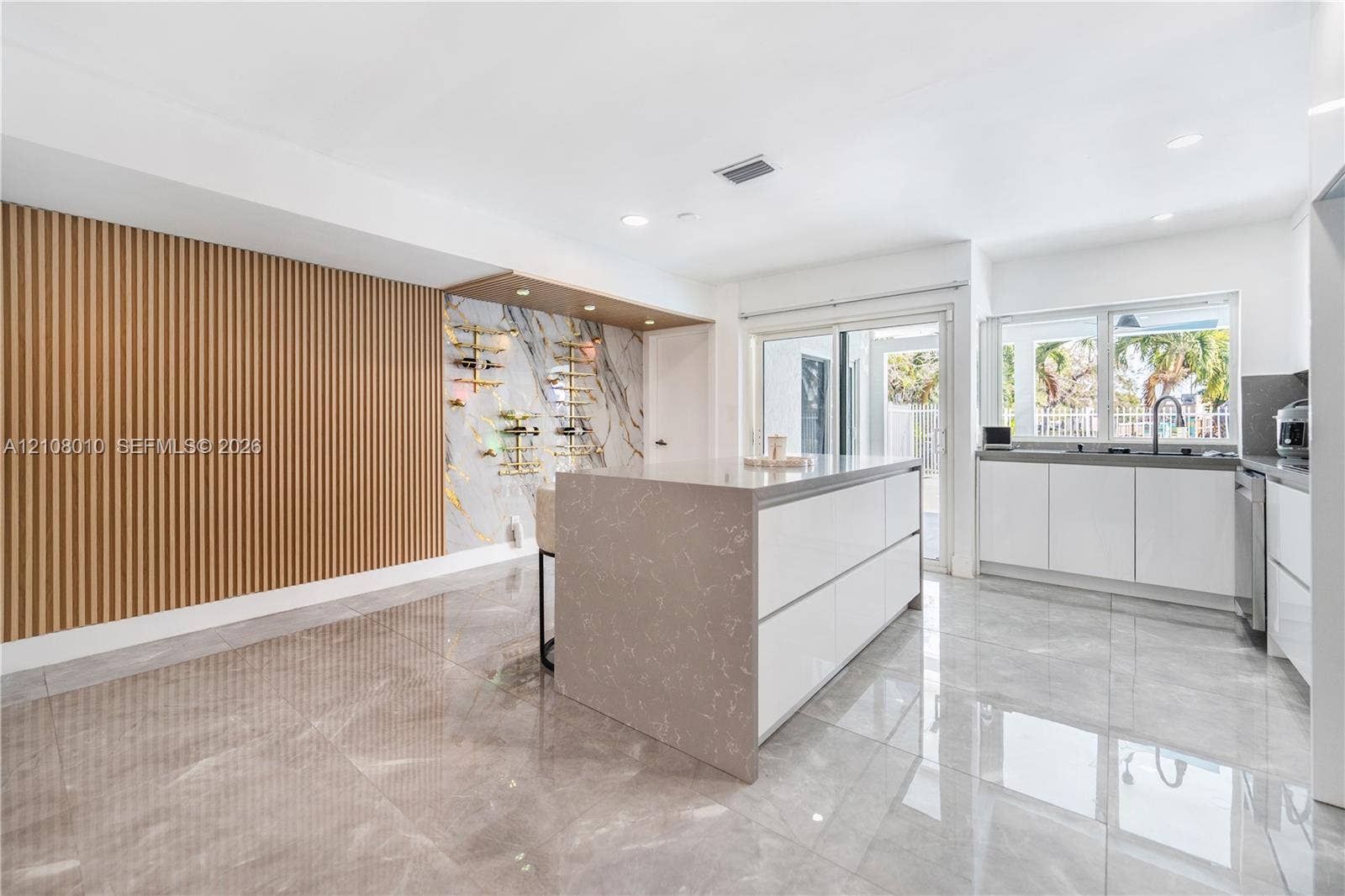 Modern kitchen featuring large-format tile flooring, a center island with solid-surface countertop, vertical wood-slat accent wall, and a custom built-in wine display with stone-finish backsplash