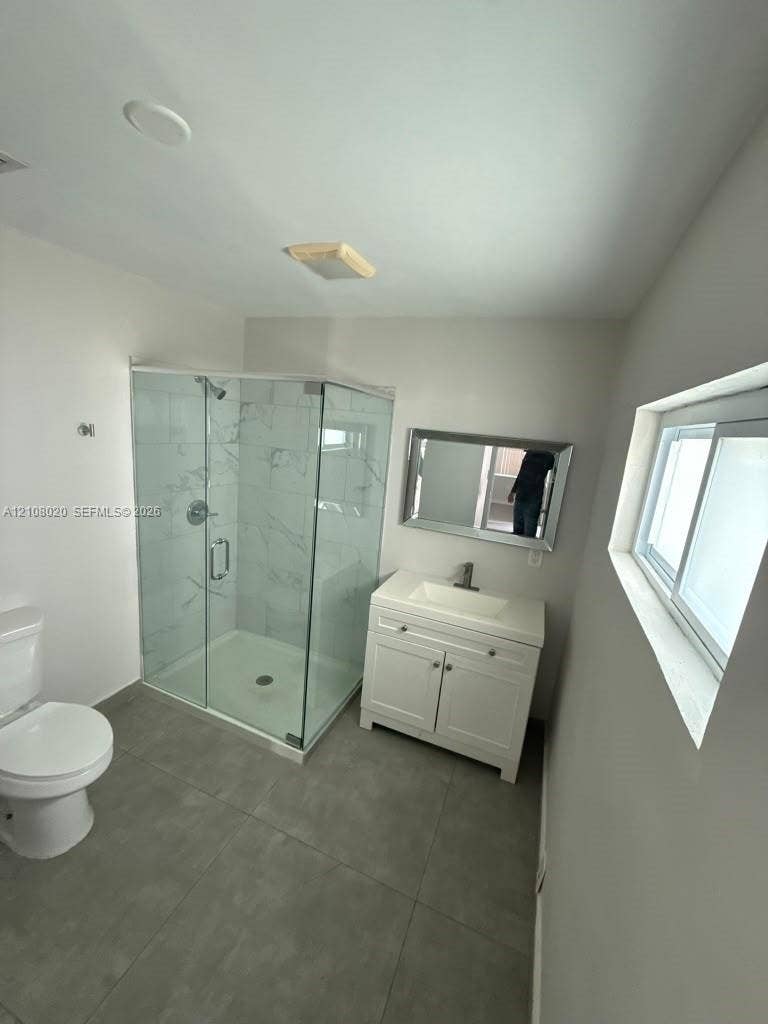 Corner shower enclosure with glass panels and patterned wall tile, white vanity cabinet with a rectangular basin, gray floor tile, and a wall-mounted mirror