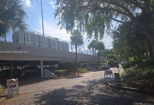 1800 NE 114th Street, #2207, Miami, FL 33181 Covered parking structure featuring stone-paver driveway, concrete canopy, chain-link perimeter fencing, and mature palm landscaping