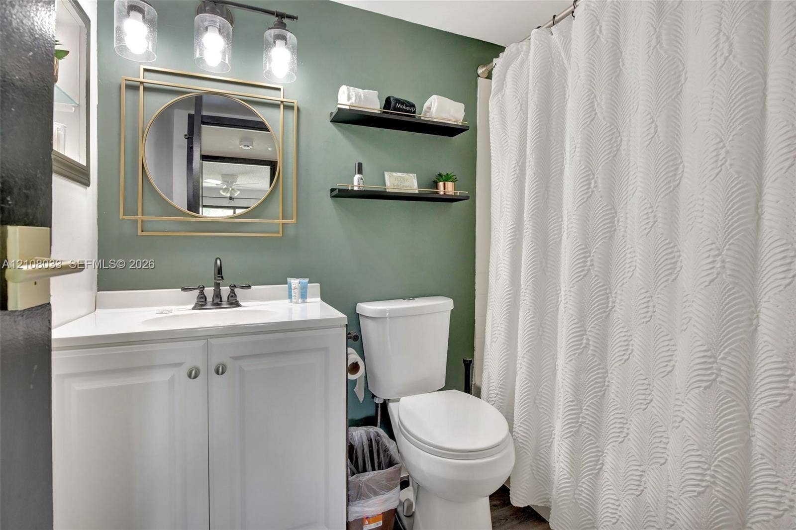 5821 Washington Street, #30, Hollywood, Hollywood, FL 33023 Bathroom featuring a white vanity cabinet, solid-surface countertop with an integrated basin, three-light fixture with glass shades, geometric gold-tone wall mirror, and dual wall-mounted floating shelves