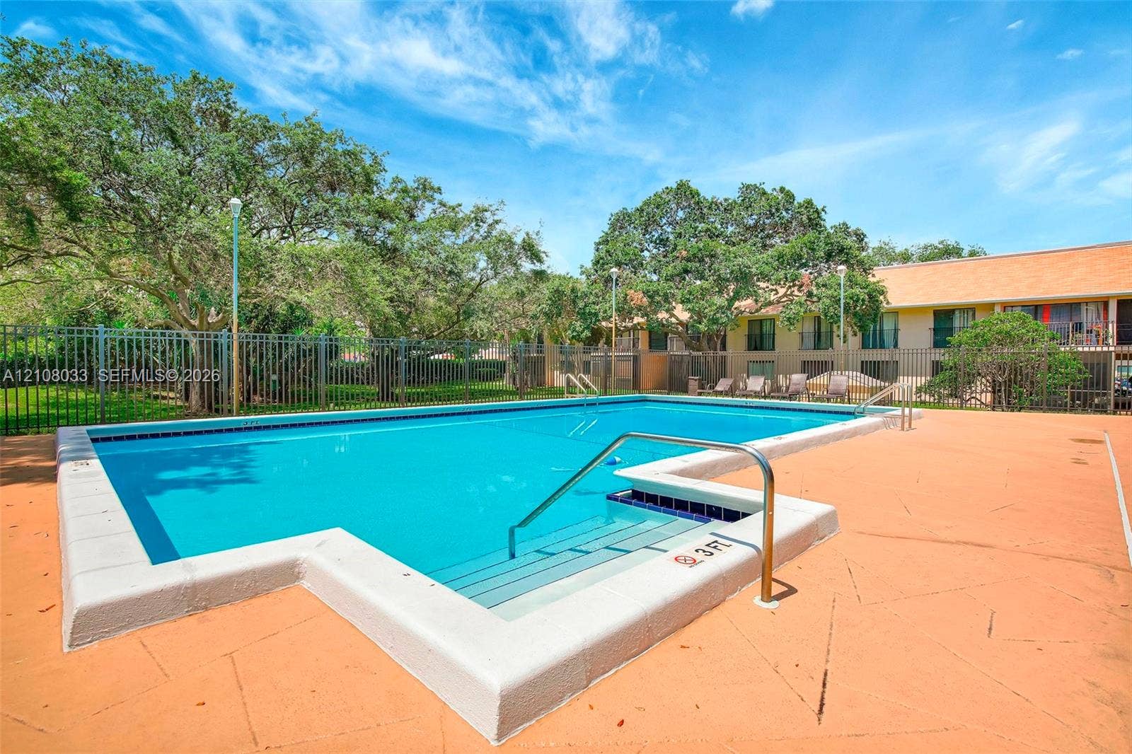 5821 Washington Street, #30, Hollywood, Hollywood, FL 33023 Rectangular swimming pool featuring a concrete deck, metal perimeter fencing, integrated steps with a metal handrail, and mature trees