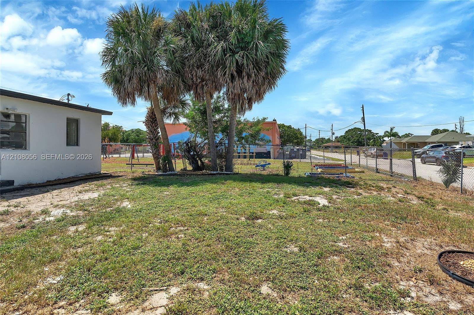 1444 W 28th Street, Riviera Beach, FL 33404 Exterior side yard featuring mature palm trees, partial chain-link fencing, and white stucco siding