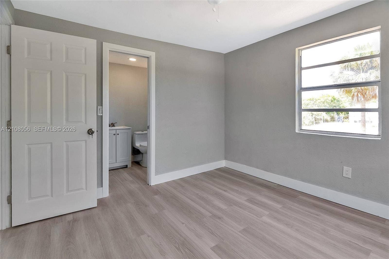 1444 W 28th Street, Riviera Beach, FL 33404 Room featuring neutral-toned walls, wood-finish flooring, a multi-pane window, and a six-panel door leading to a bathroom with a vanity cabinet and toilet