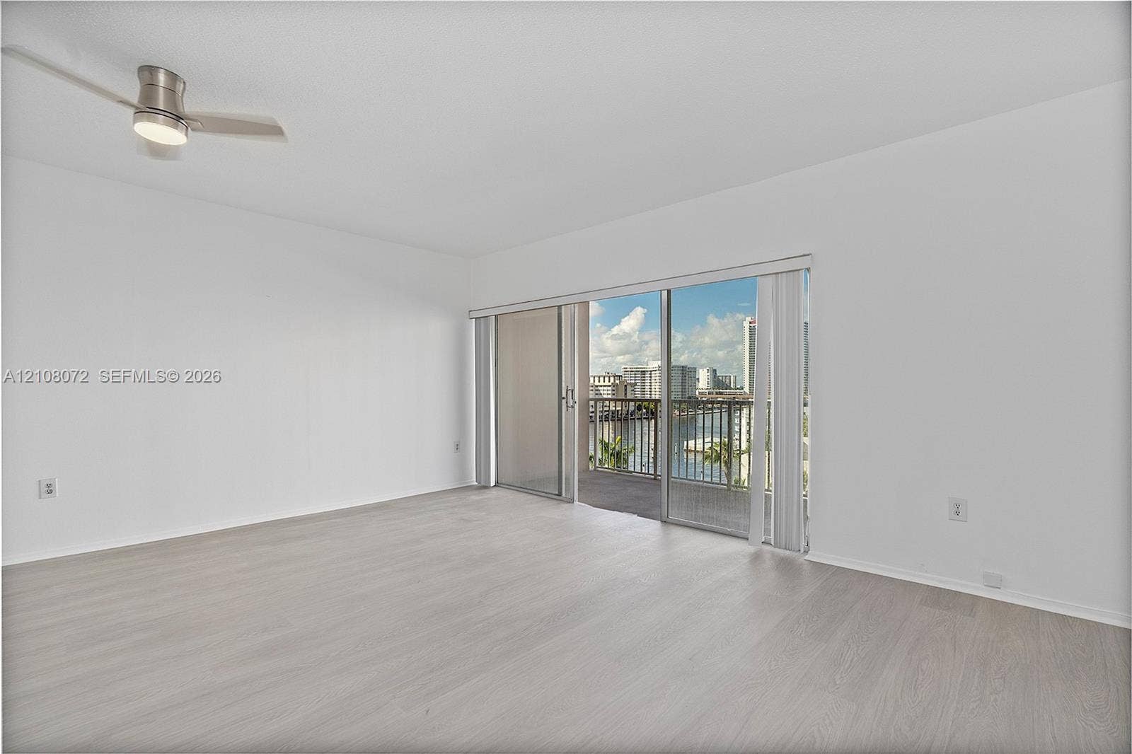 Neutral interior space featuring wood-finish flooring, a ceiling-mounted light fixture with fan, and a large sliding glass door providing access to an exterior balcony