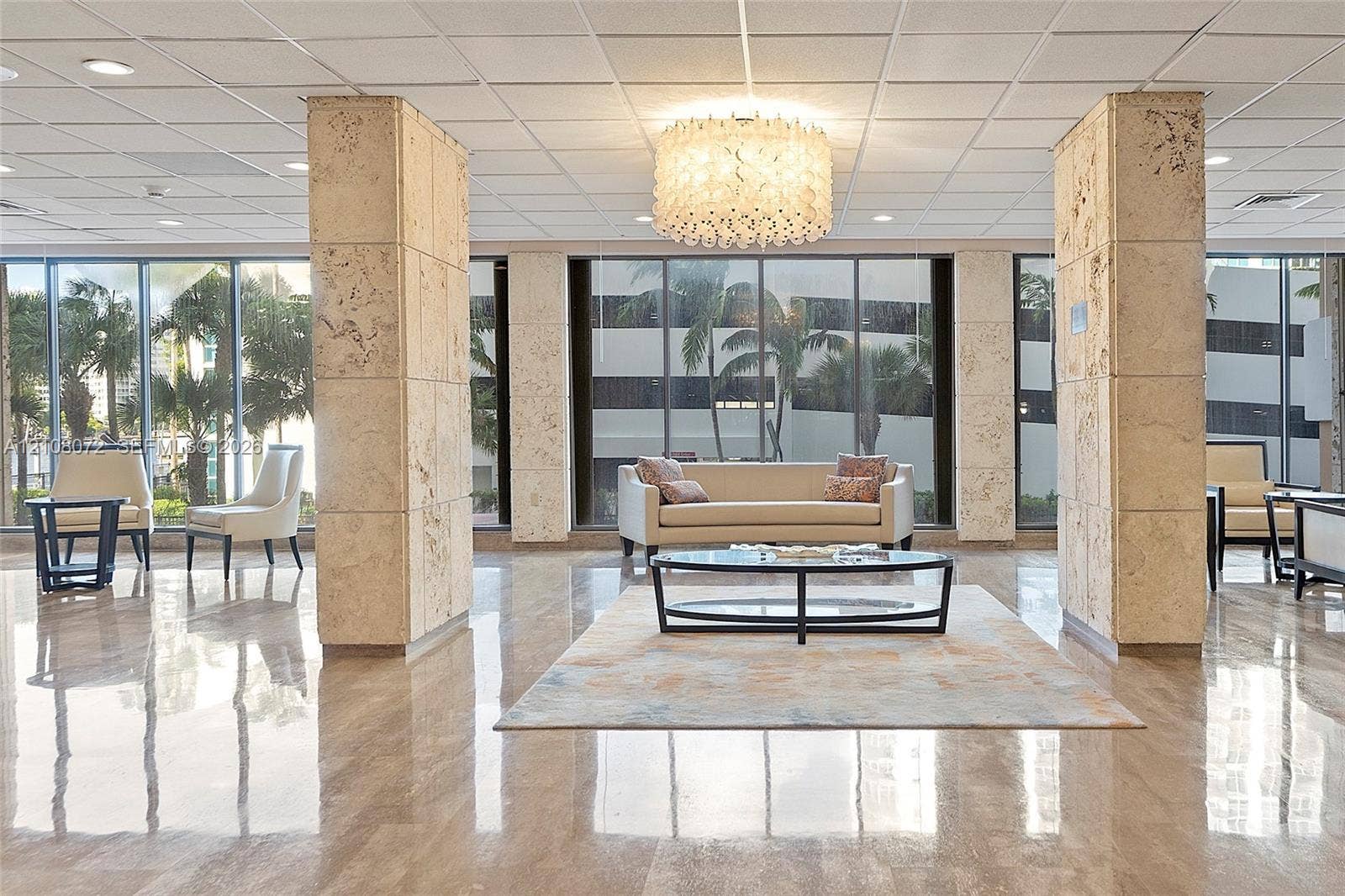 Spacious lobby featuring polished stone flooring, floor-to-ceiling windows, architectural stone-clad support pillars, a multi-tiered circular chandelier, and a drop ceiling