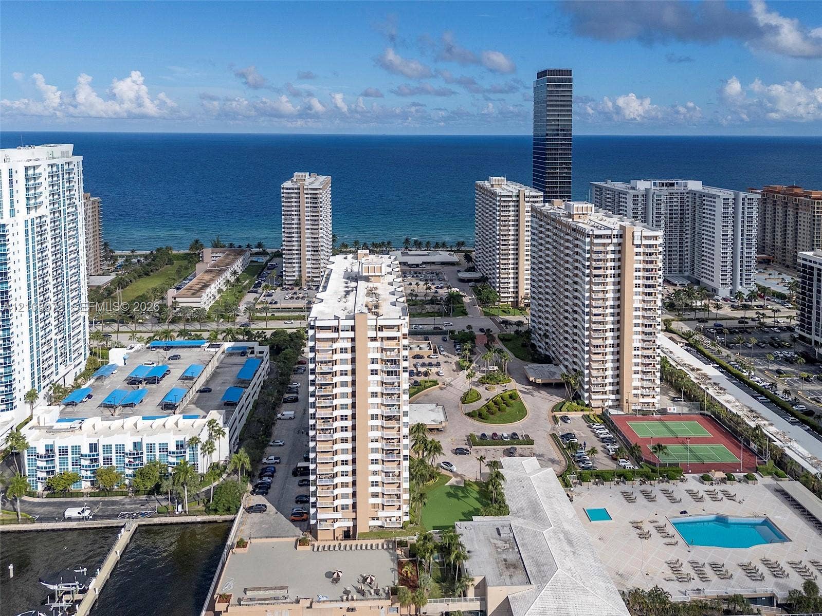 Aerial perspective featuring waterfront residential buildings, outdoor swimming pool, hard-surfaced tennis courts, and landscaped green space