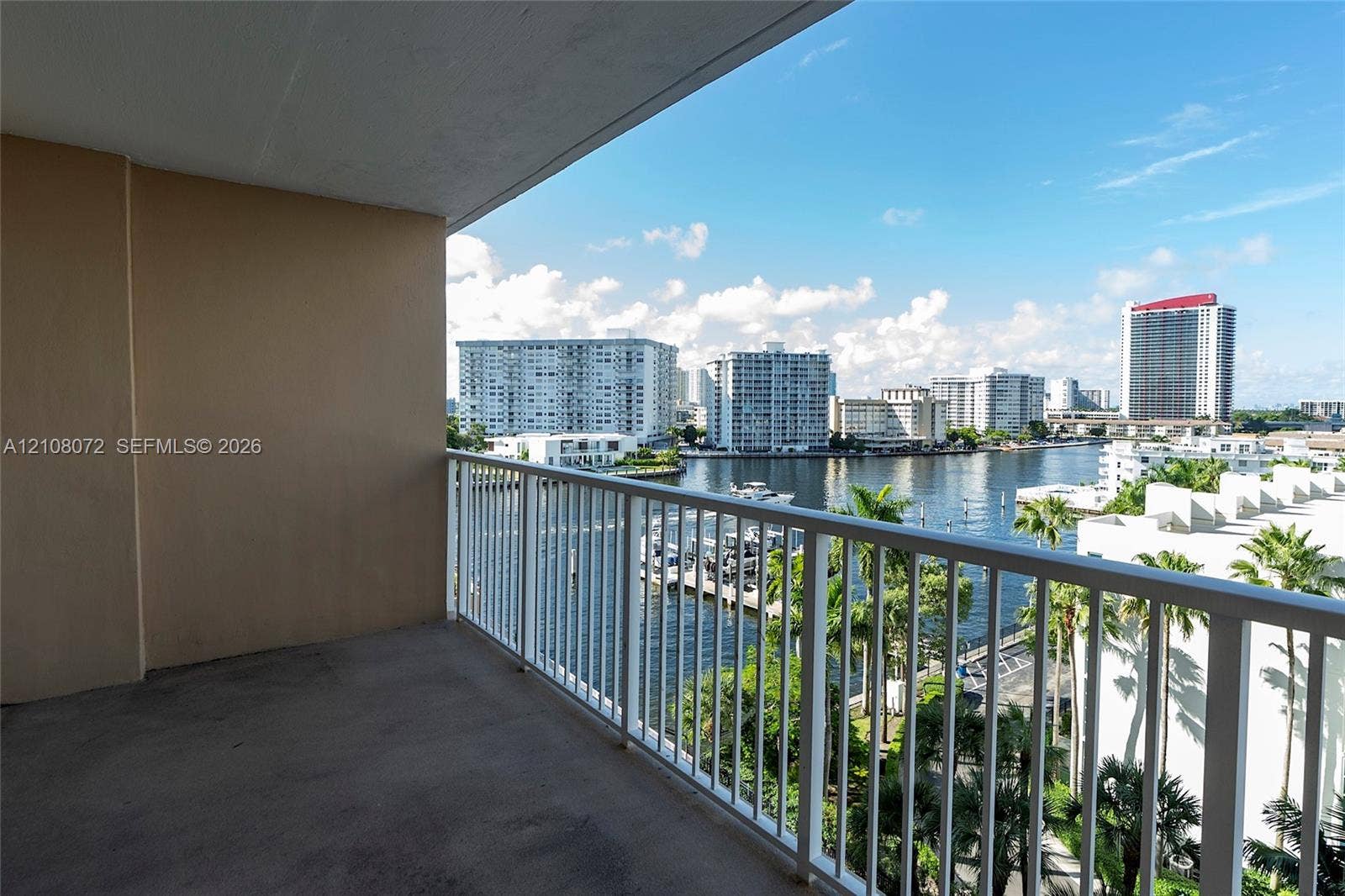 Covered balcony featuring concrete flooring and a white metal railing, overlooking a waterway with urban scenery