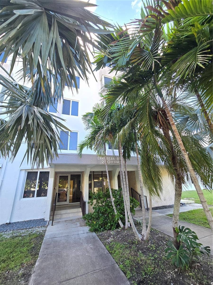 250 180th Drive, #202, Sunny Isles Beach, Sunny Isles Beach, FL 33160 Multi-story exterior featuring light-toned facade, concrete walkway, and mature palm landscaping