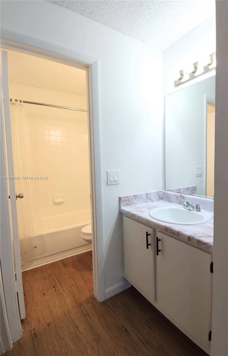 4401 W Mcnab Road, Pompano Beach, FL 33069 Bathroom featuring a vanity with white cabinetry, matte black hardware, and a stone-patterned countertop with an integrated basin, paired with wood-finish flooring and a wall-mounted light fixture above the mirror