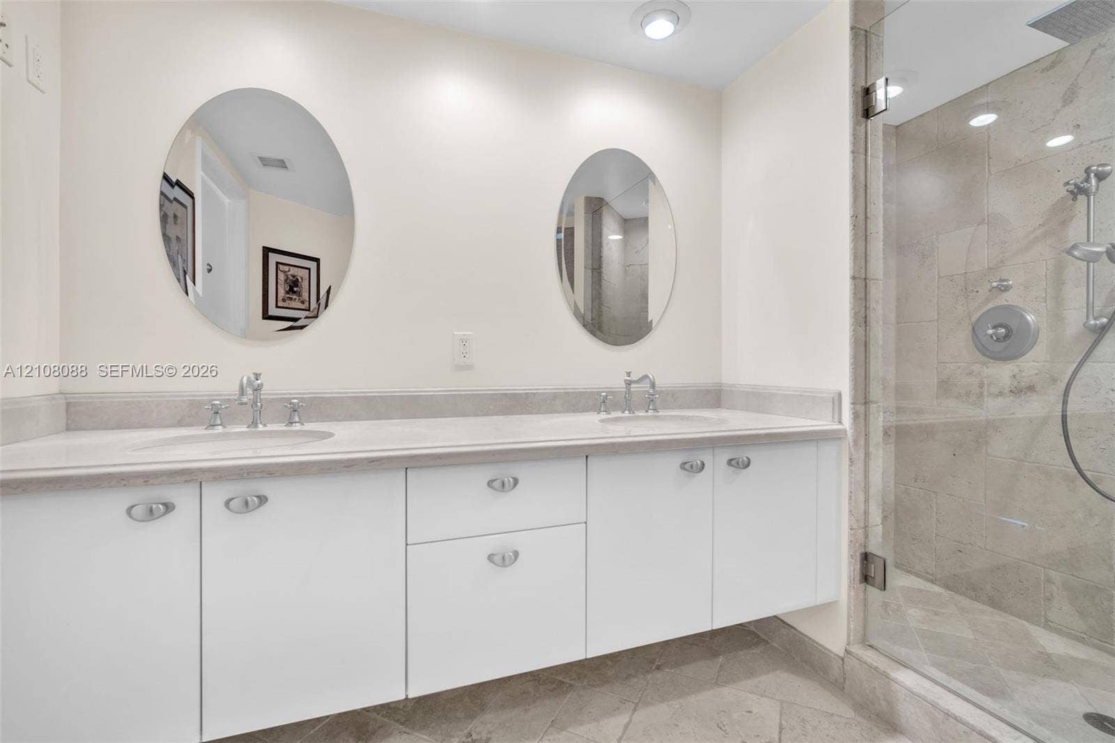 333 Las Olas Way, #2601, Fort Lauderdale, FL 33301 Dual-sink vanity with white cabinetry, light-toned stone-finish countertop, dual oval mirrors, and a glass-enclosed shower featuring stone wall tiles