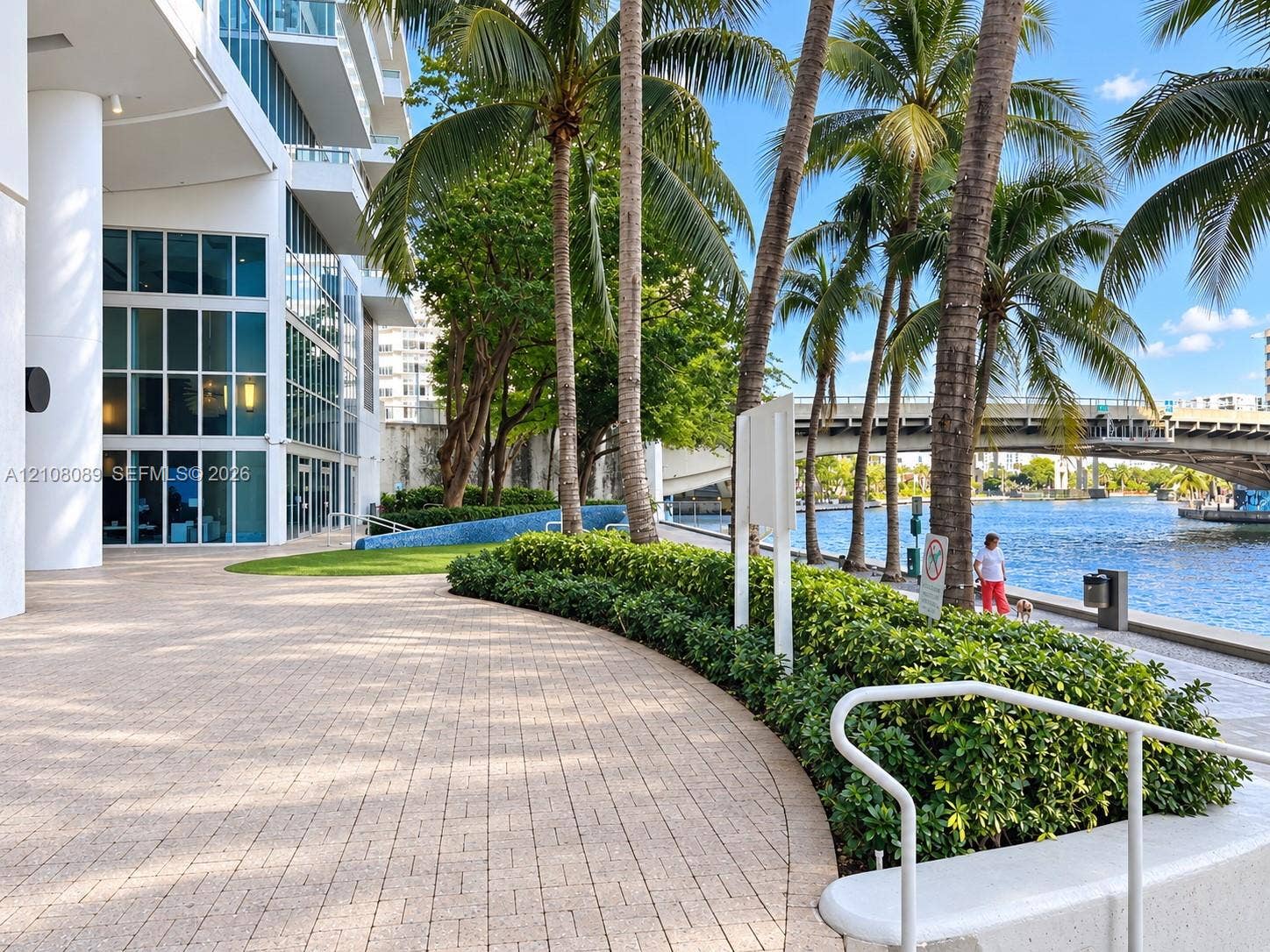 185 SW 7th Street, #3608, Miami, FL 33130 Waterfront walkway featuring paver hardscaping, mature palm trees, manicured hedges, and floor-to-ceiling glass building facade