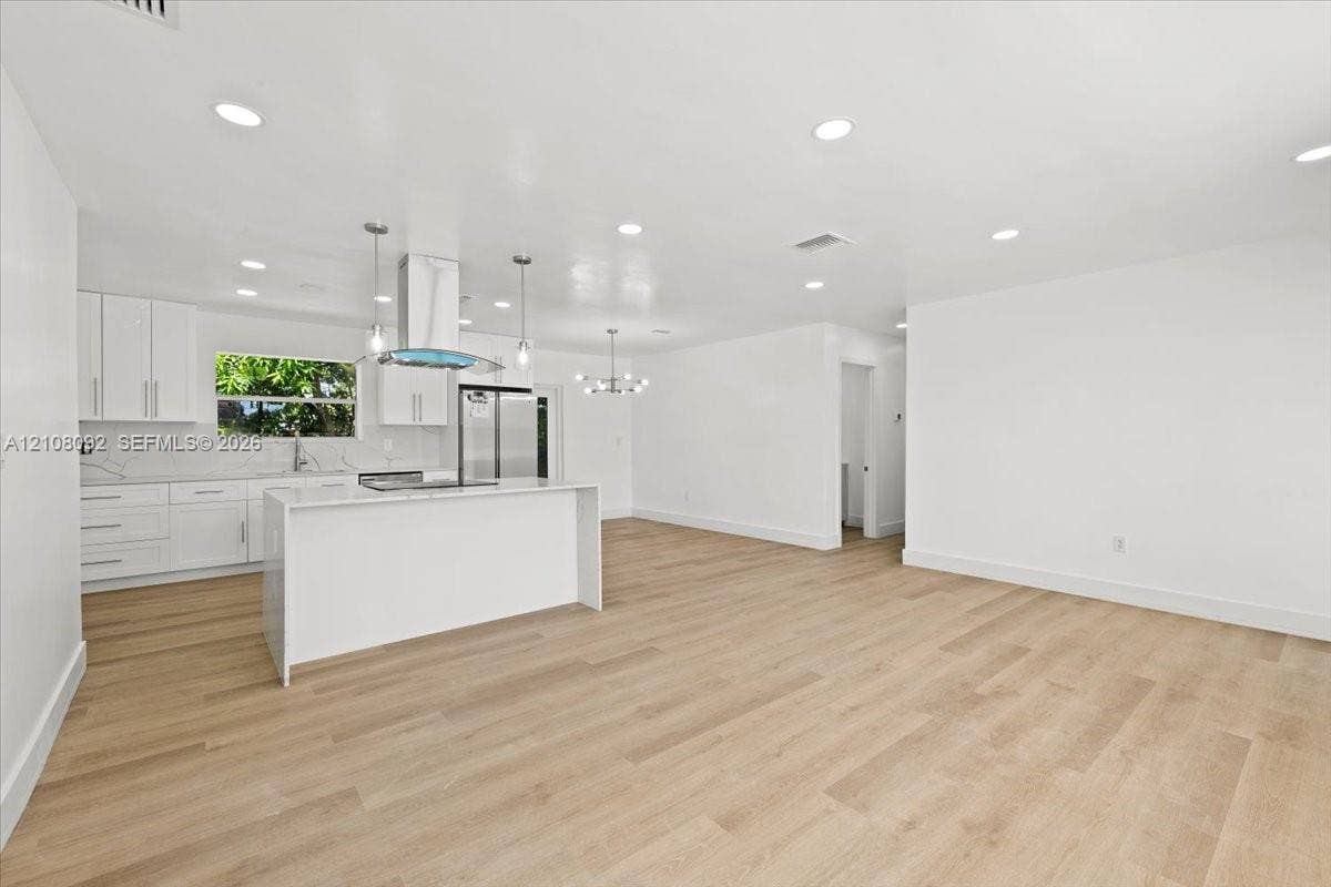 Open-concept interior featuring light wood-finish flooring, recessed lighting, a central island with stone-finish waterfall countertop, stainless steel appliances, and white shaker-style cabinetry