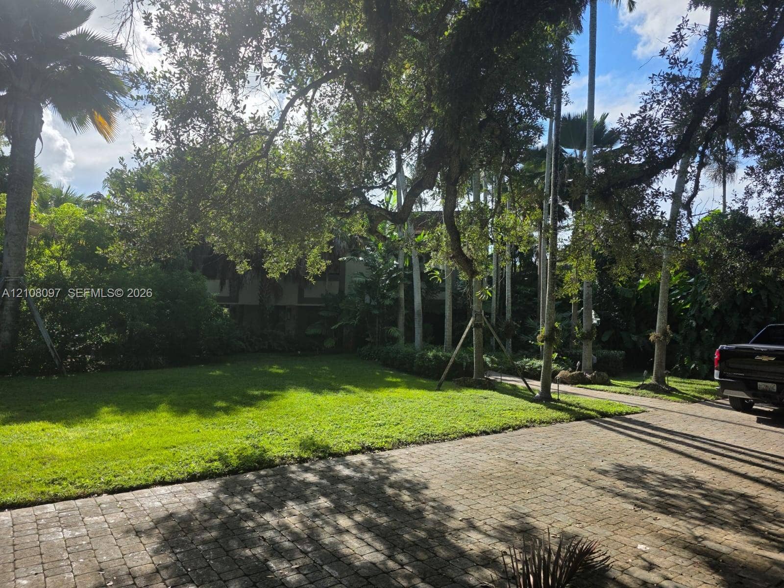 Expansive grounds featuring a paved brick driveway, mature canopy trees, manicured lawn, and established tropical landscaping