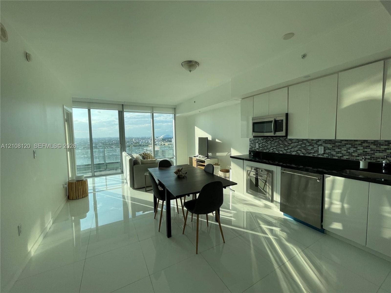92 SW 3rd Street, #2307, Miami, FL 33130 Open-concept floor plan featuring high-gloss tile flooring, floor-to-ceiling glass paneling, white flat-panel cabinetry, stainless steel appliances, and a glass-tile mosaic backsplash