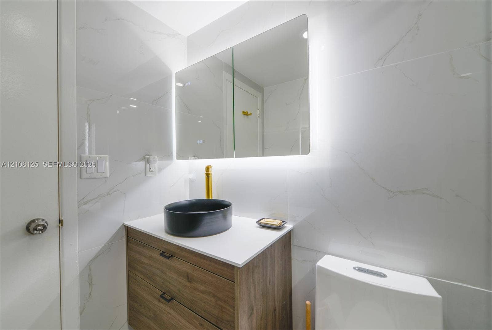 1985 S Ocean Drive, #8M, Hallandale Beach, FL 33009 Modern vanity with wood-finish cabinetry, white countertop, black vessel sink, and brass faucet, complemented by wall-to-wall light-toned tiling and an integrated backlit medicine cabinet