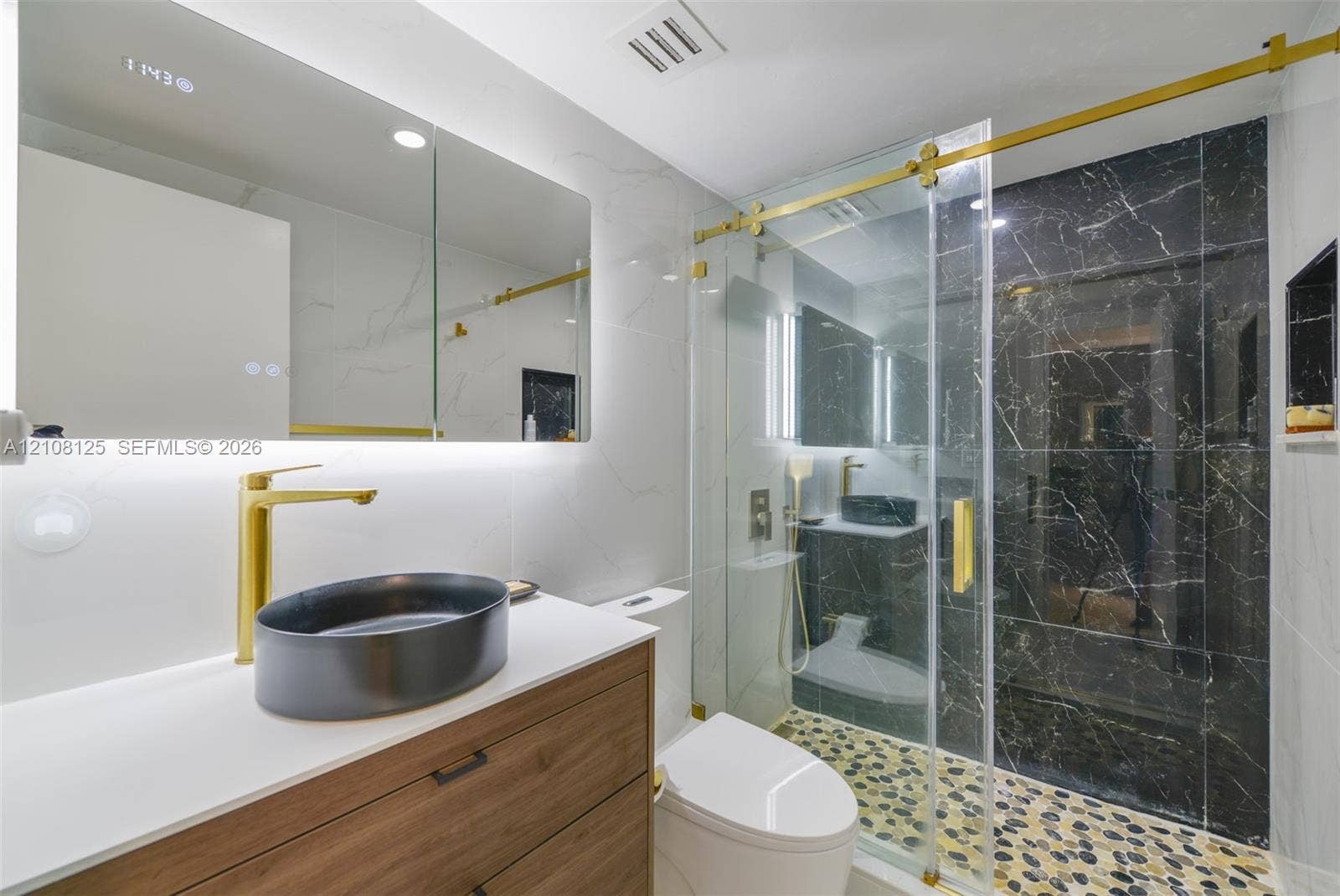 1985 S Ocean Drive, #8M, Hallandale Beach, FL 33009 Bathroom featuring a wood-finish vanity with a quartz countertop, oval vessel sink, gold-toned hardware, and a walk-in shower with stone-tile flooring and dark accent tiling