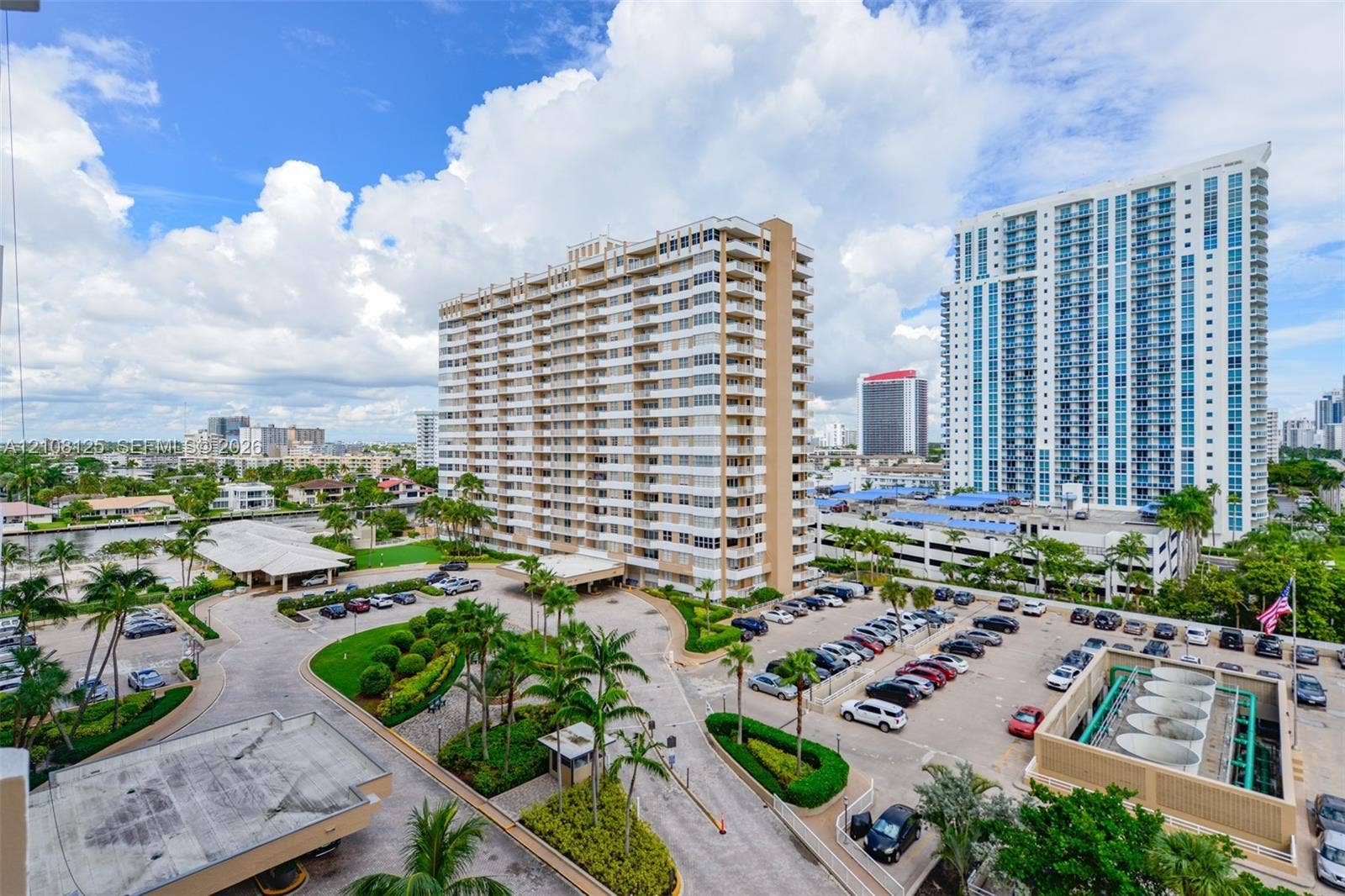1985 S Ocean Drive, #8M, Hallandale Beach, FL 33009 High-rise building with a tiered concrete facade, expansive surface parking, paved circular driveway with a covered entrance, landscaped grounds with mature palm trees, and canal frontage
