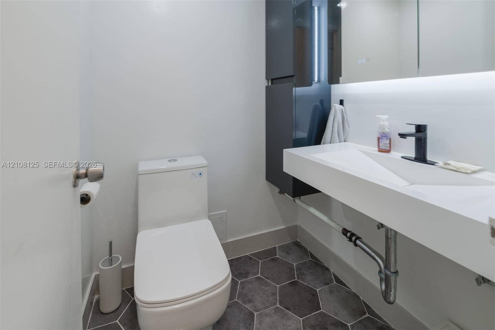 1985 S Ocean Drive, #8M, Hallandale Beach, FL 33009 Modern powder room featuring a wall-mounted floating sink, matte black faucet, integrated storage cabinetry, back-lit mirror, and charcoal hexagon floor tile