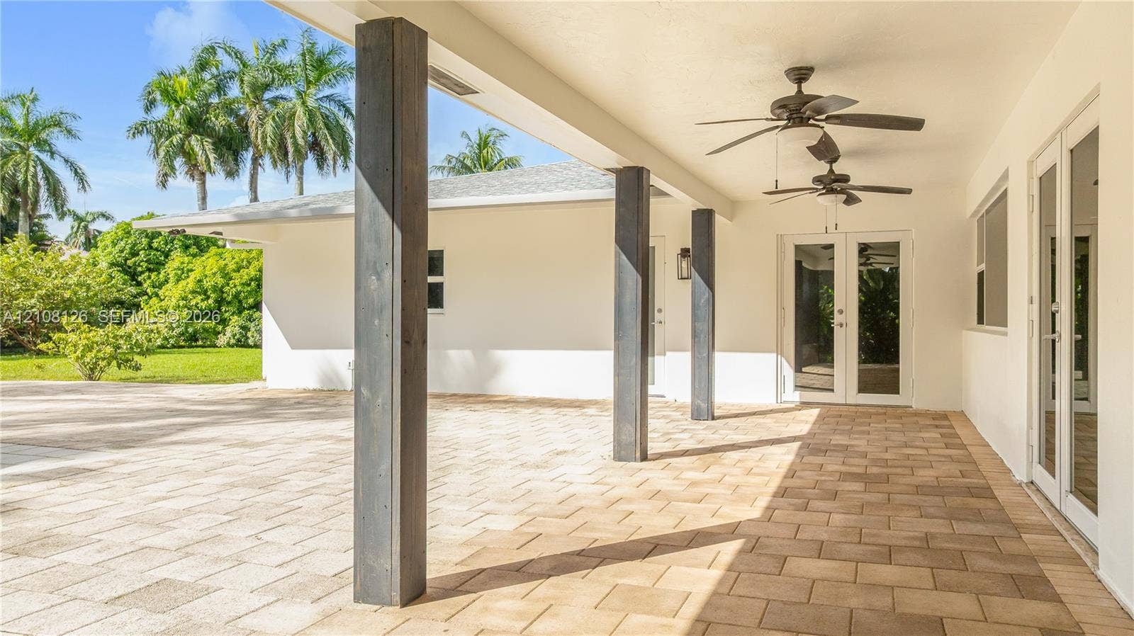 11700 NW 4th Street, Plantation, Plantation, FL 33325 Covered outdoor patio featuring light-toned paver flooring, dark wood-finish structural support columns, dual ceiling fans, and double glass-paned doors
