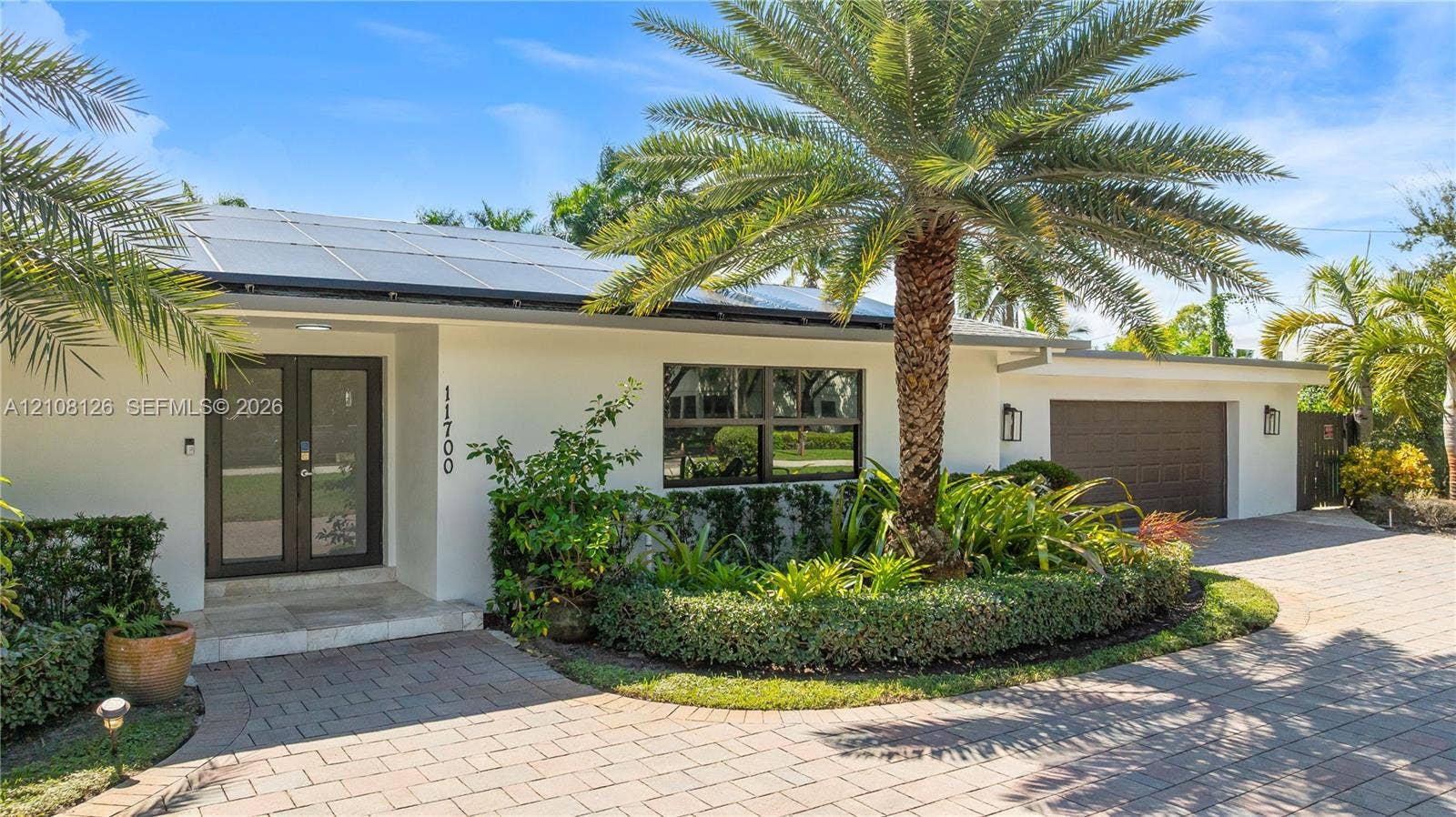 11700 NW 4th Street, Plantation, Plantation, FL 33325 Single-story exterior featuring a light-toned stucco facade, rooftop solar panels, a paved brick driveway, a double-entry glass door, and integrated outdoor wall lighting