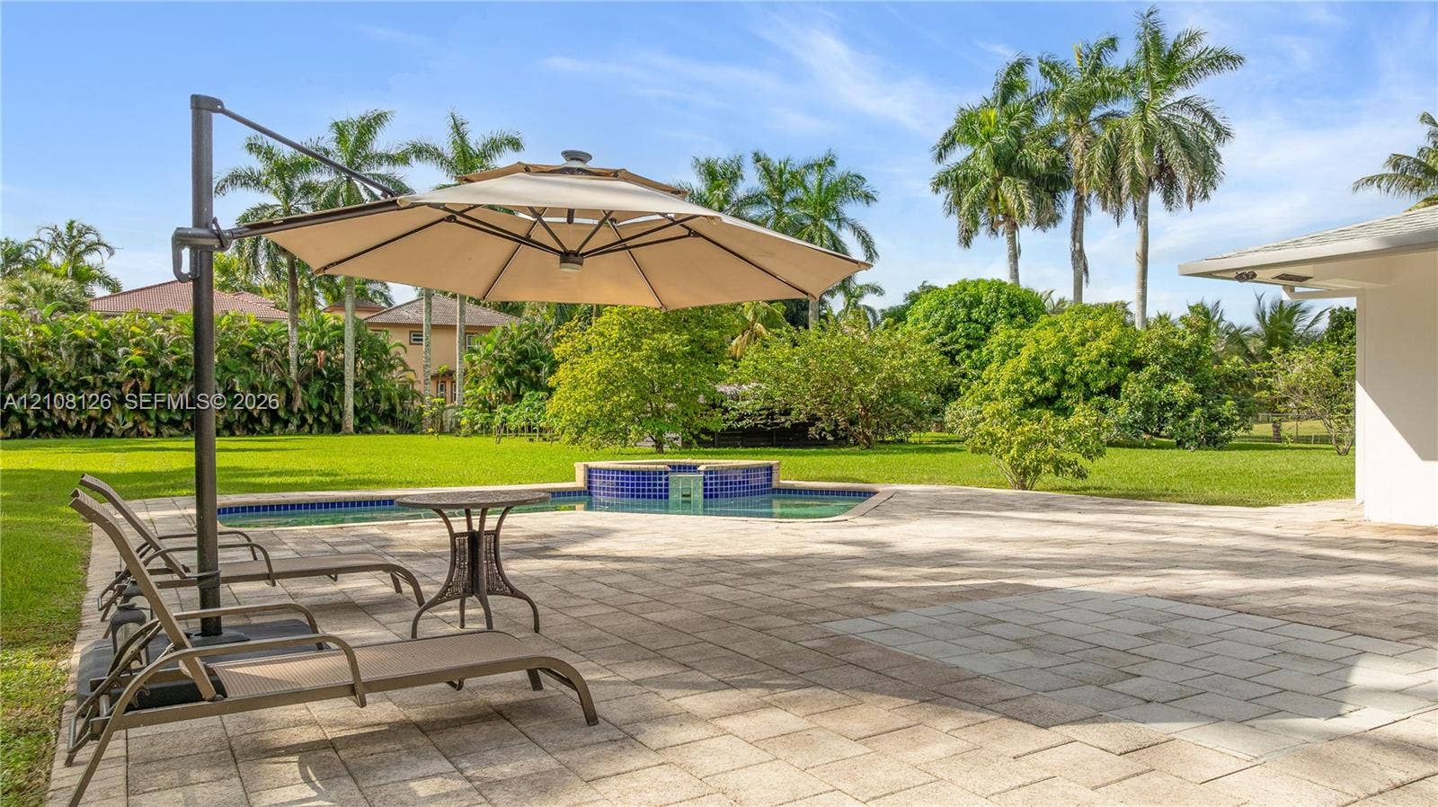 11700 NW 4th Street, Plantation, Plantation, FL 33325 Expansive backyard featuring a stone-paver patio, swimming pool with a blue-tiled raised spillover spa, manicured lawn, and mature palm trees