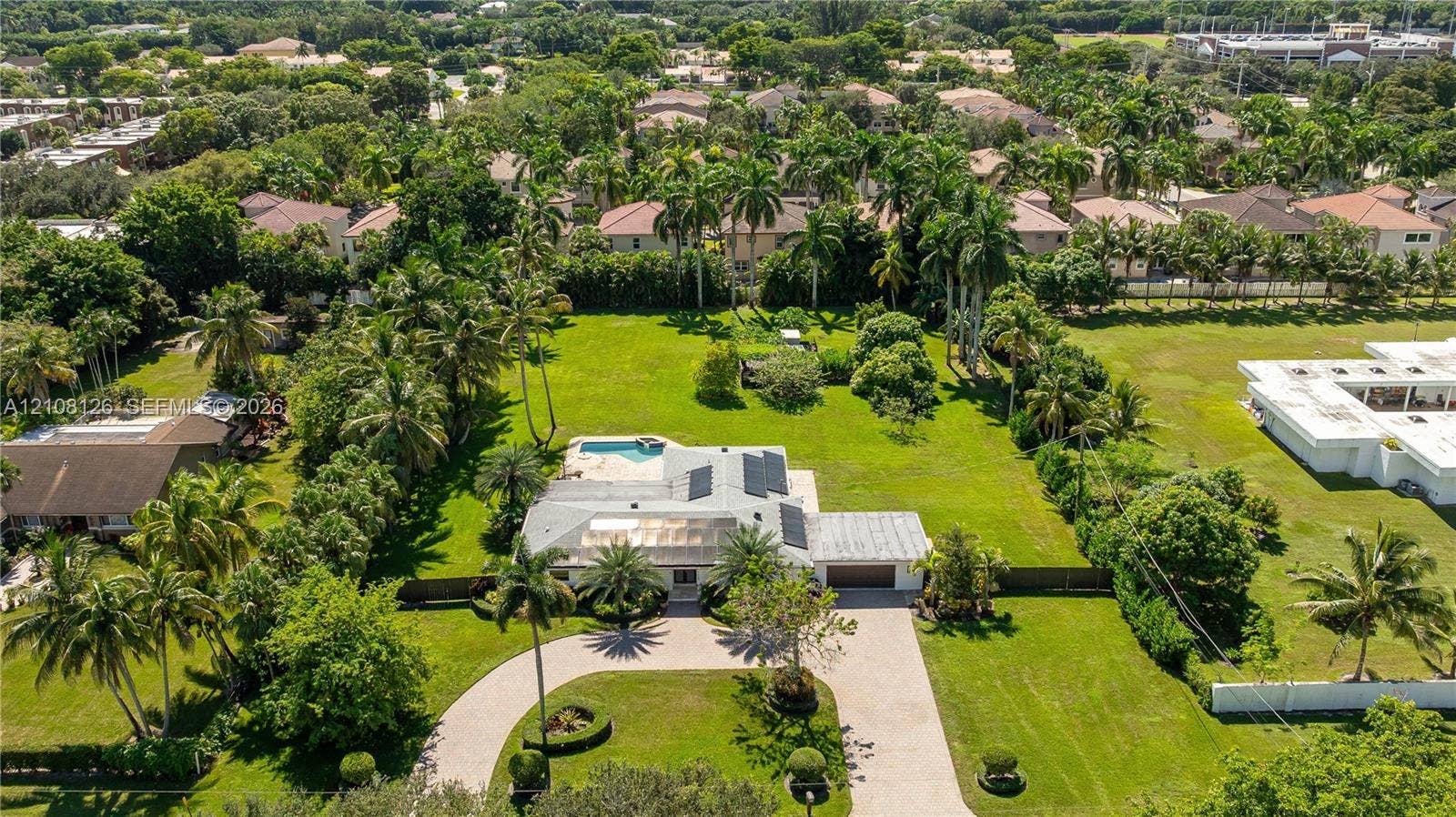 11700 NW 4th Street, Plantation, Plantation, FL 33325 Aerial view of a sprawling property featuring a circular paved driveway, a multi-car garage, solar roof panels, a screened-in swimming pool, and expansive manicured grounds