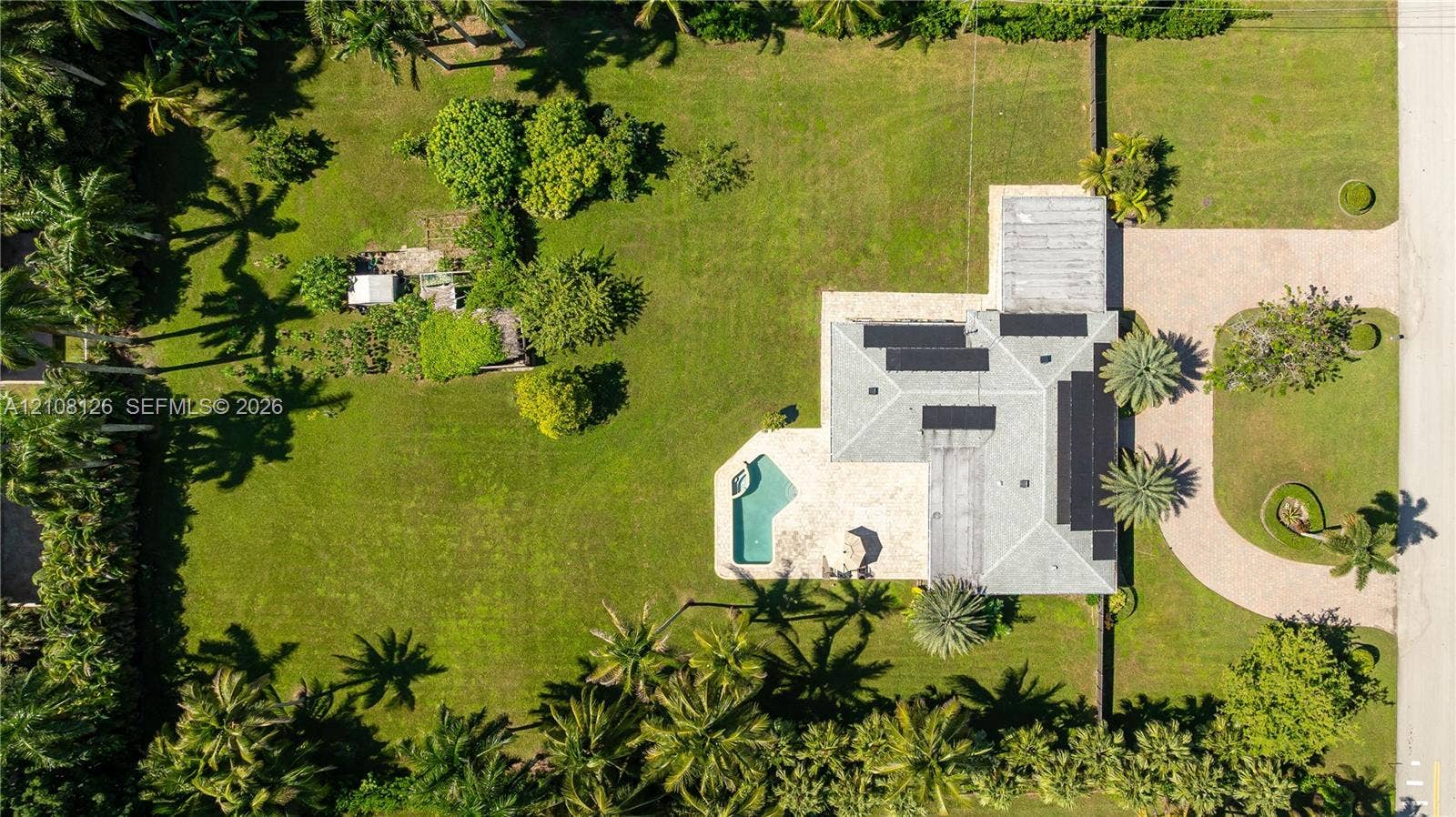 11700 NW 4th Street, Plantation, Plantation, FL 33325 Aerial perspective featuring an expansive landscaped lot, paved circular driveway, integrated swimming pool with stone-surfaced patio, and a shingled roof with solar panels