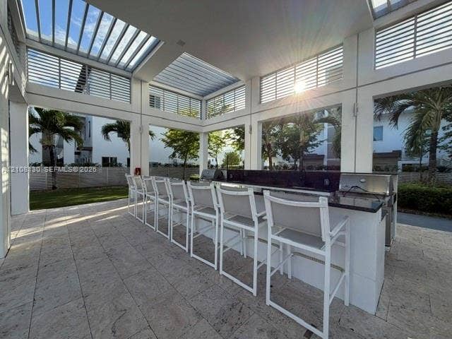 Covered outdoor kitchen featuring stone tile flooring, a built-in counter with a dark stone surface, stainless steel grills, and a slatted pergola roof structure