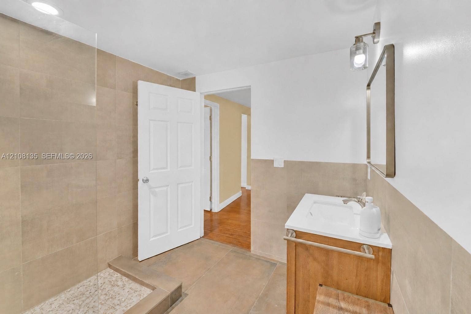 Bathroom featuring a walk-in shower with large-format tile surround, tiled shower floor, glass partition, wood-finish vanity, and wall-mounted lighting