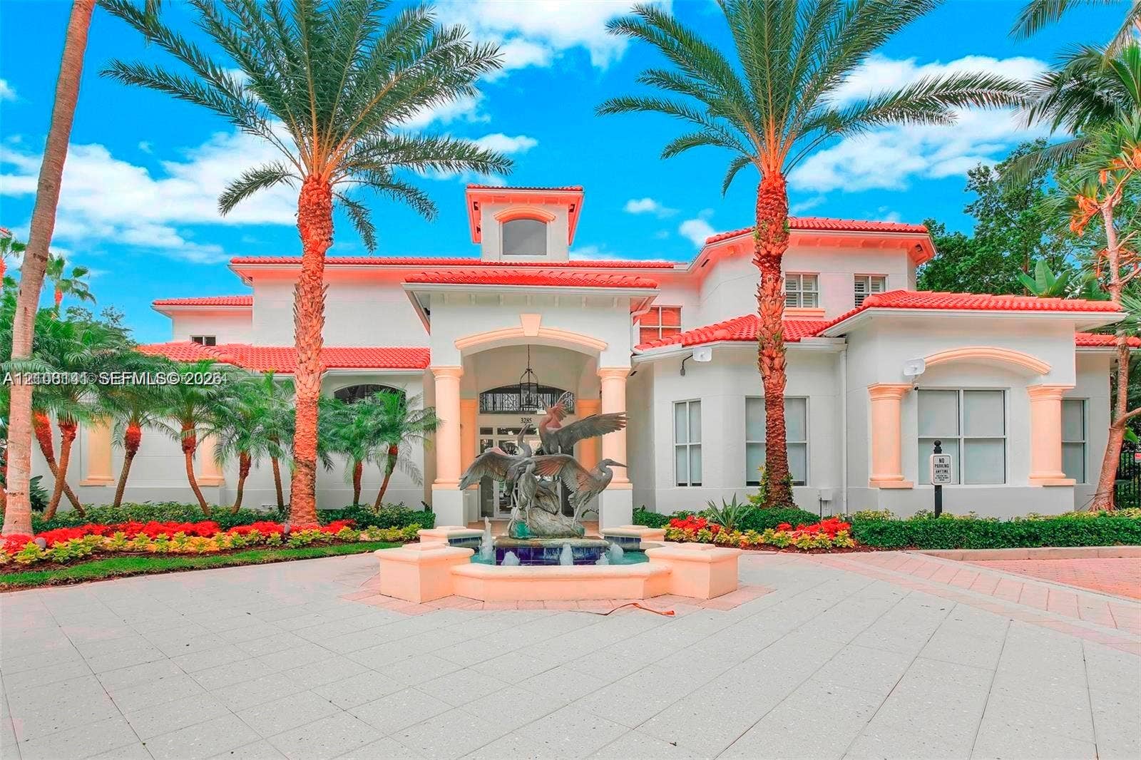 3135 NE 184th Street, #2205, Aventura, FL 33160 Mediterranean-style exterior featuring a red tile roof, stucco facade, covered entryway with columns, a central decorative fountain, and palm-lined landscaping