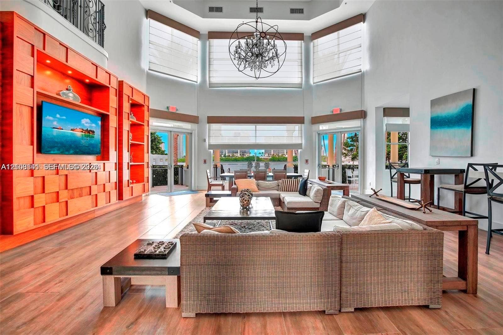 3135 NE 184th Street, #2205, Aventura, FL 33160 Double-height ceilings with large multi-panel windows, custom floor-to-ceiling wood-finish media cabinetry, wood-finish flooring, and a large decorative chandelier
