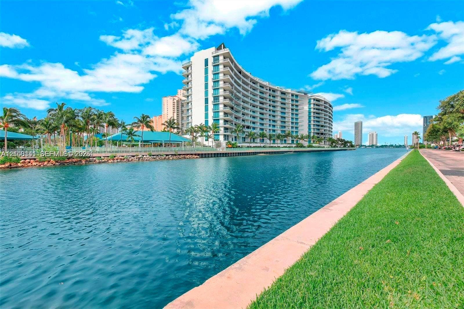 3135 NE 184th Street, #2205, Aventura, FL 33160 Curved mid-rise architectural facade with expansive balconies, situated on a waterfront lot featuring stone bulkhead, manicured grass landscaping, and established palm trees