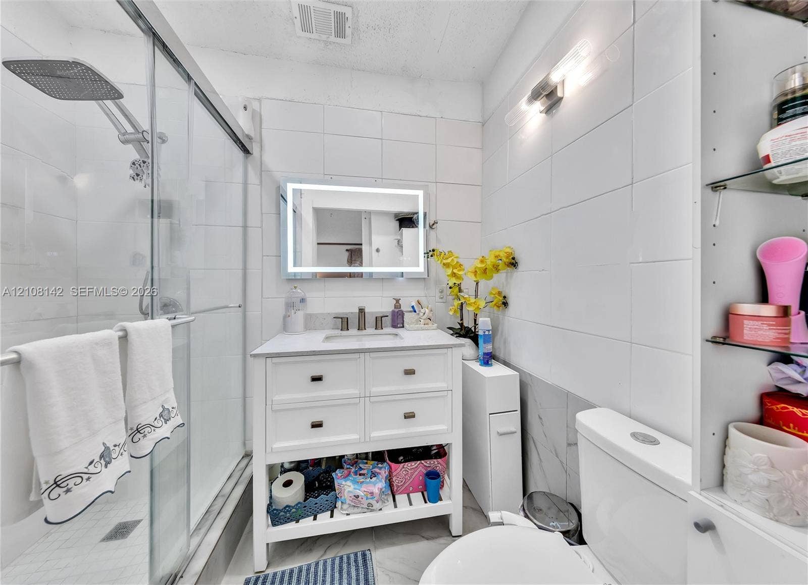 Bathroom featuring a walk-in shower with glass enclosure, white square tile wall surround, vanity with integrated basin, LED-backlit mirror, and built-in shelving
