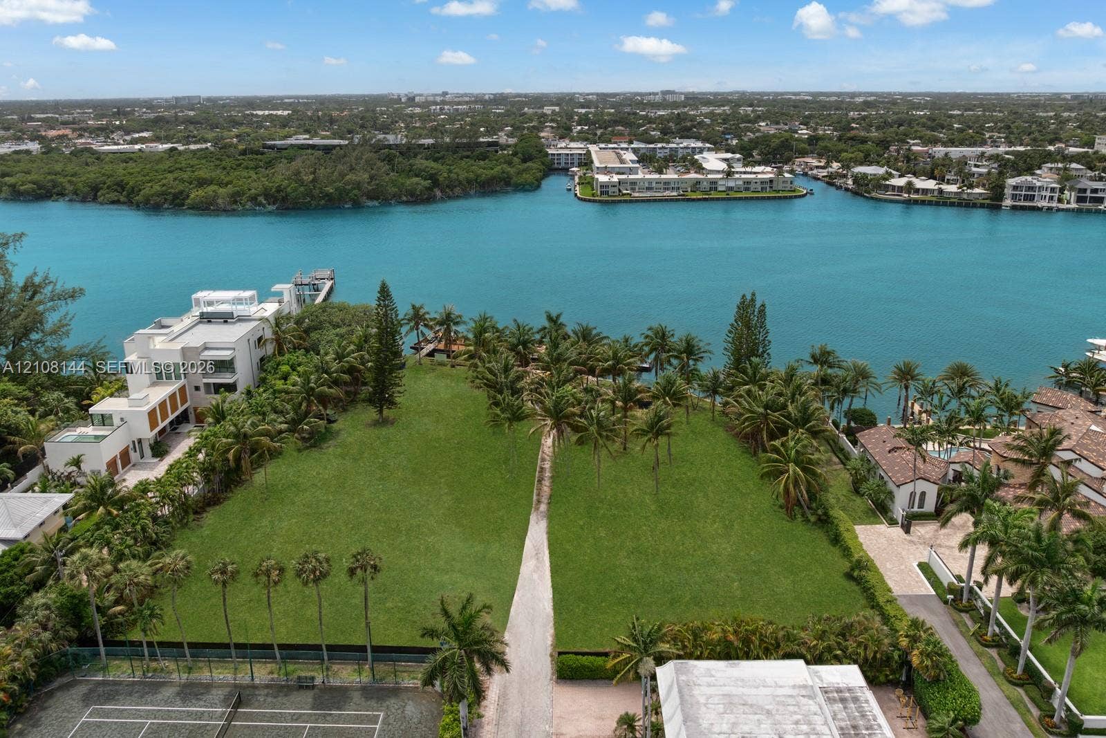 Waterfront lot featuring direct canal access, expansive manicured lawn, mature palm trees, and a hard-surface tennis court