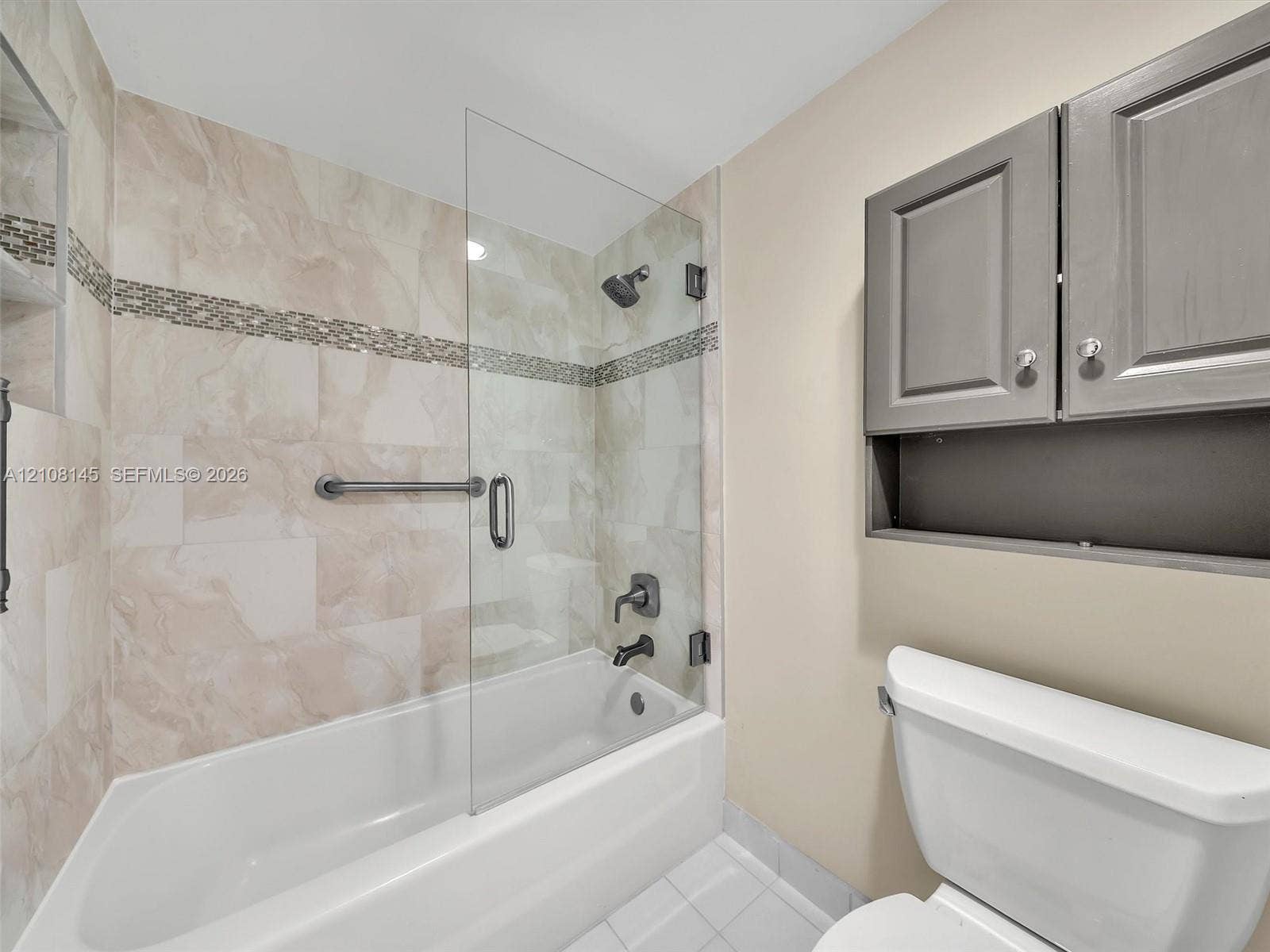1020 Citrus Way, #101, Delray Beach, FL 33445 Bathtub featuring a glass shower panel, neutral tile surround with decorative mosaic accent, matte black wall-mounted fixtures, and built-in grey cabinetry